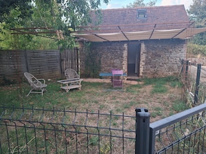 A stone house with a covered patio, a wooden fence, and outdoor furniture. - Gîte/Studio sleeps 2 with enclosed garden (pets welcome) and large shower. (Loubressac)