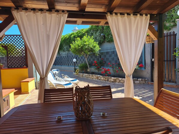 A covered patio with a wooden table, two lounge chairs, and a view of a garden with a fence and a tree.
