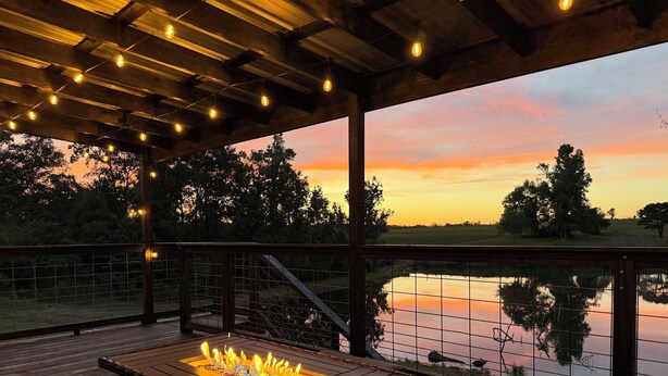 Une terrasse en bois dotée d’un foyer et offrant une vue sur un coucher de soleil se reflétant sur une étendue d’eau.