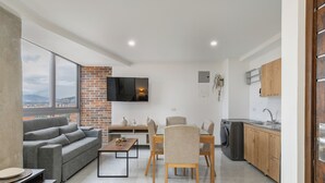 Una sala moderna con un sofá gris, una mesa de comedor con sillas, un televisor montado en la pared y una zona de cocina con armarios de madera y una lavadora.