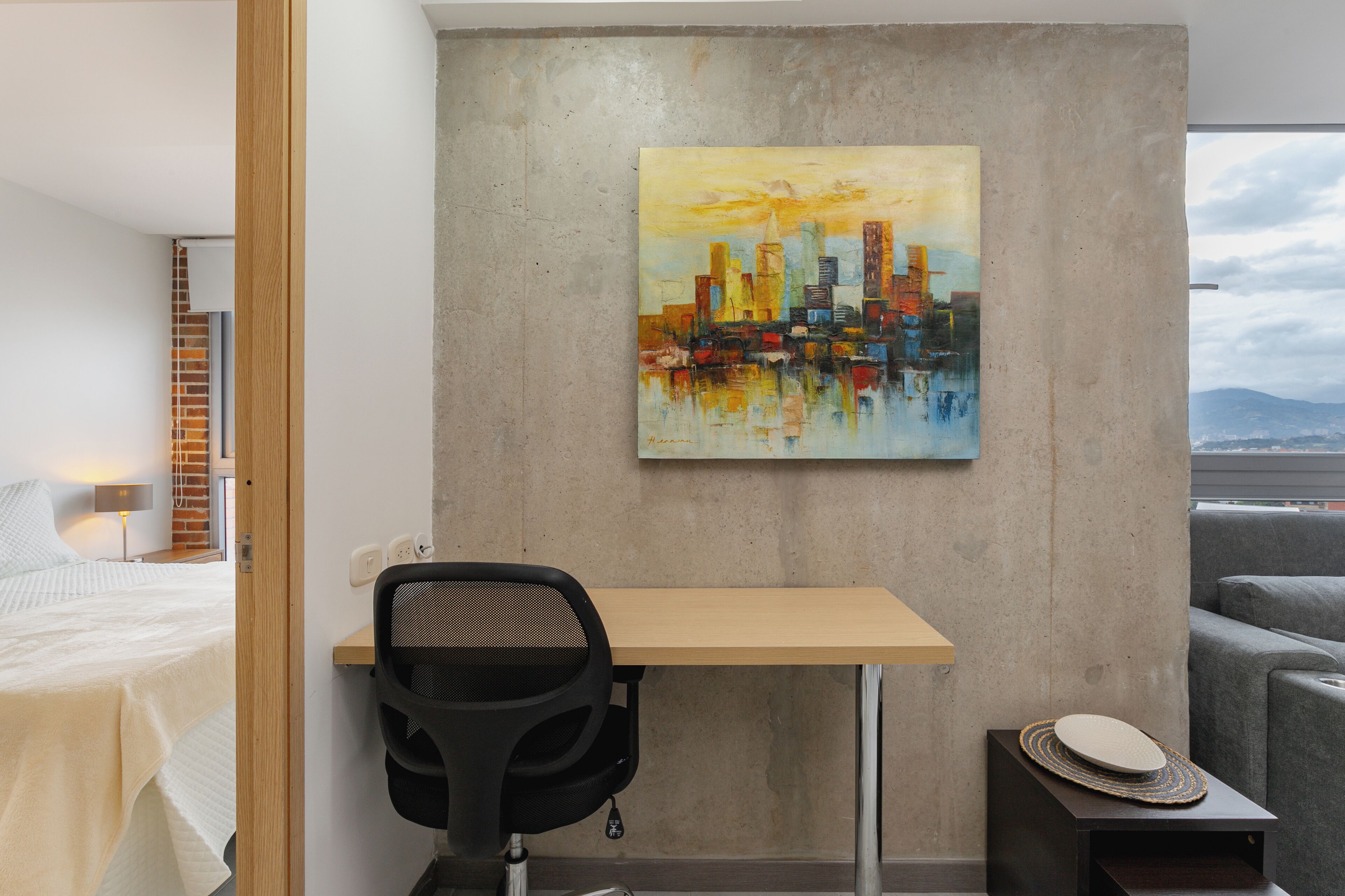 A modern hotel room with a bed, a desk, a chair, a painting of a cityscape, and a view of the mountains.