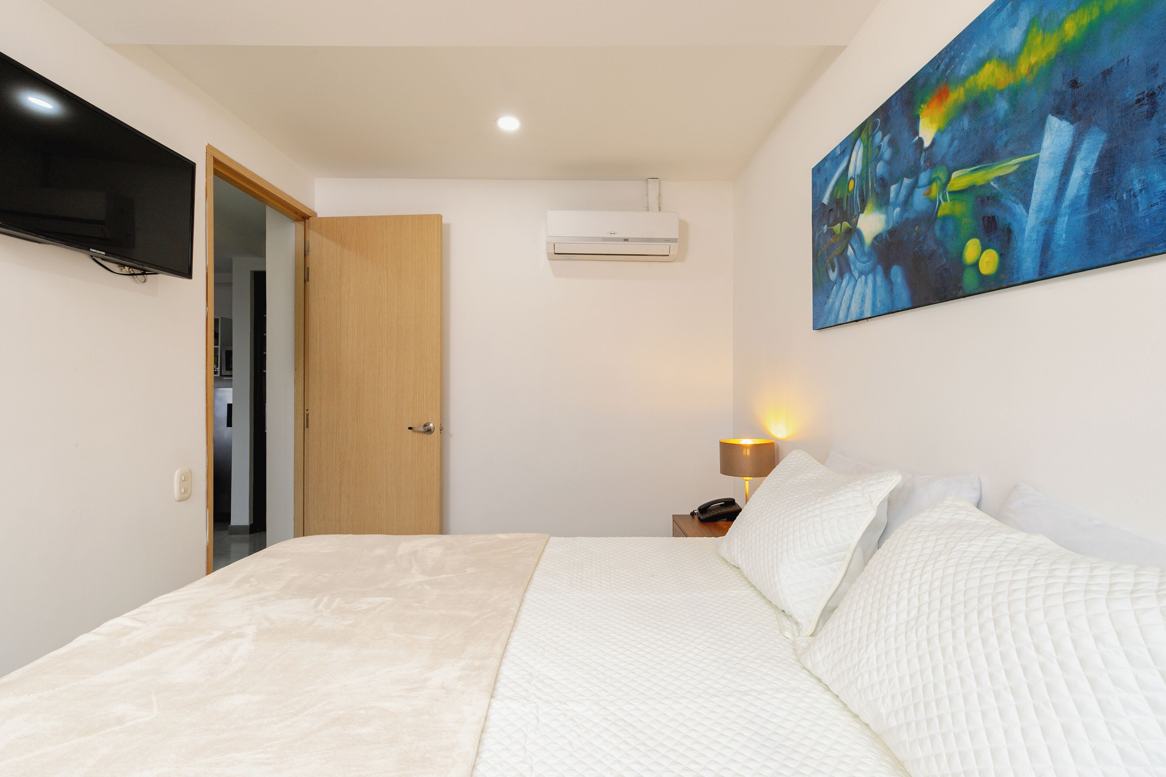 Un dormitorio con cama, televisión de pantalla plana, aire acondicionado, un cuadro y una lámpara de noche.
