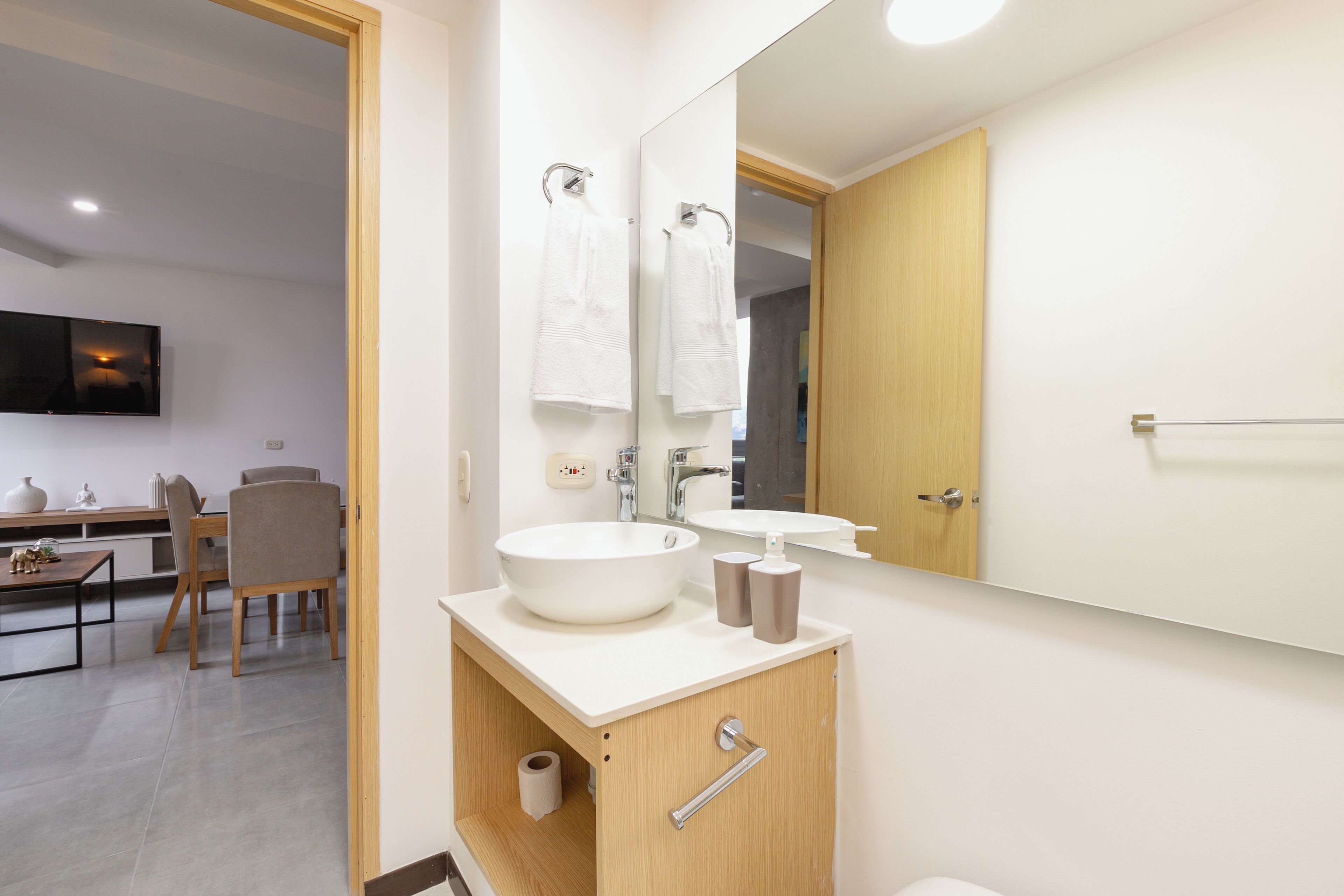 Une salle de bain moderne avec un grand miroir, un lavabo et des toilettes. La pièce adjacente est dotée d’une porte en bois et d’une petite table avec des chaises.