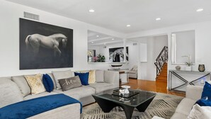 Ein modernes Wohnzimmer mit einem großen Gemälde eines Pferdes, einem grauen Sofa mit gemusterten Kissen, einem schwarzen Couchtisch und einer Treppe, die in eine andere Ebene führt.
