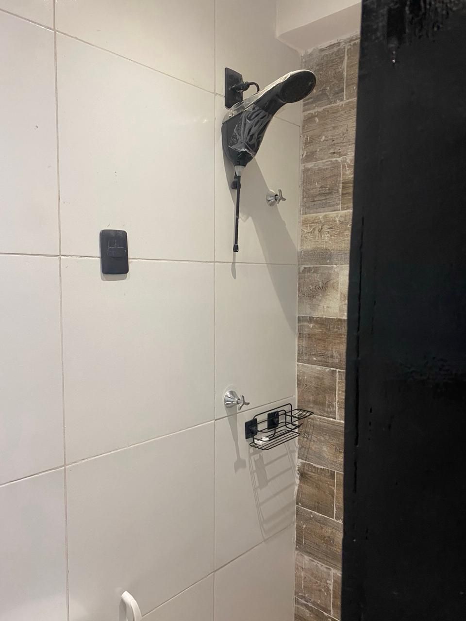 Un baño con paredes de azulejos blancos, una ducha con grifo negro y un estante pequeño con artículos de aseo.