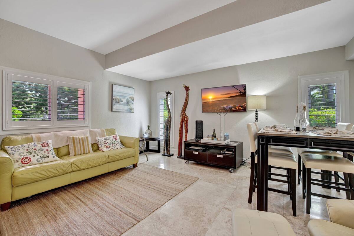 A living room with a yellow sofa, a dining table with chairs, a giraffe statue, and a painting of a sunset.