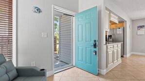 A modern living room with a grey sofa, a teal door leading to a kitchen, and a small outdoor area visible through the door. - Charming 3BR Beach House w/ Yard & Grill ☀️ (North Myrtle Beach)