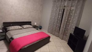 A bedroom with a bed, a nightstand, a TV stand, and a window with floral curtains. - Safe Refuge (Macerata Campania)