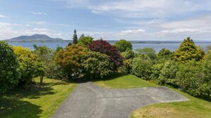 A paved driveway leading to a grassy area with trees and a view of a body of water and distant mountains. - Acacia Bay  - Lake and Mountain Views (Taupō)
