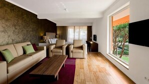 A modern living room with a sofa, armchairs, a coffee table, a TV, and a large window with a view. - Vilamoura Garden Hotel (Vilamoura)
