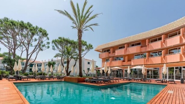 A swimming pool with wooden deck, lounge chairs, umbrellas, and a multi-story building with balconies. - Vilamoura Garden Hotel (Vilamoura)