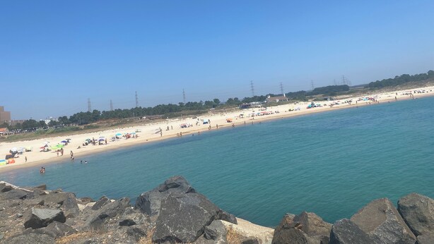 Une plage avec une eau d’un bleu limpide, un premier plan rocheux et une rive sablonneuse parsemée de parasols et de personnes.