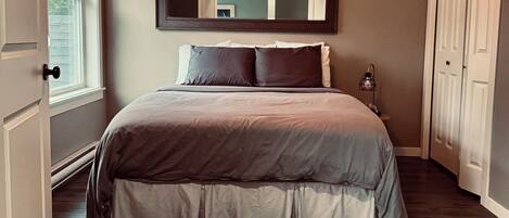 A bedroom with a large bed, a mirror above the bed, a window with blinds, and a door.