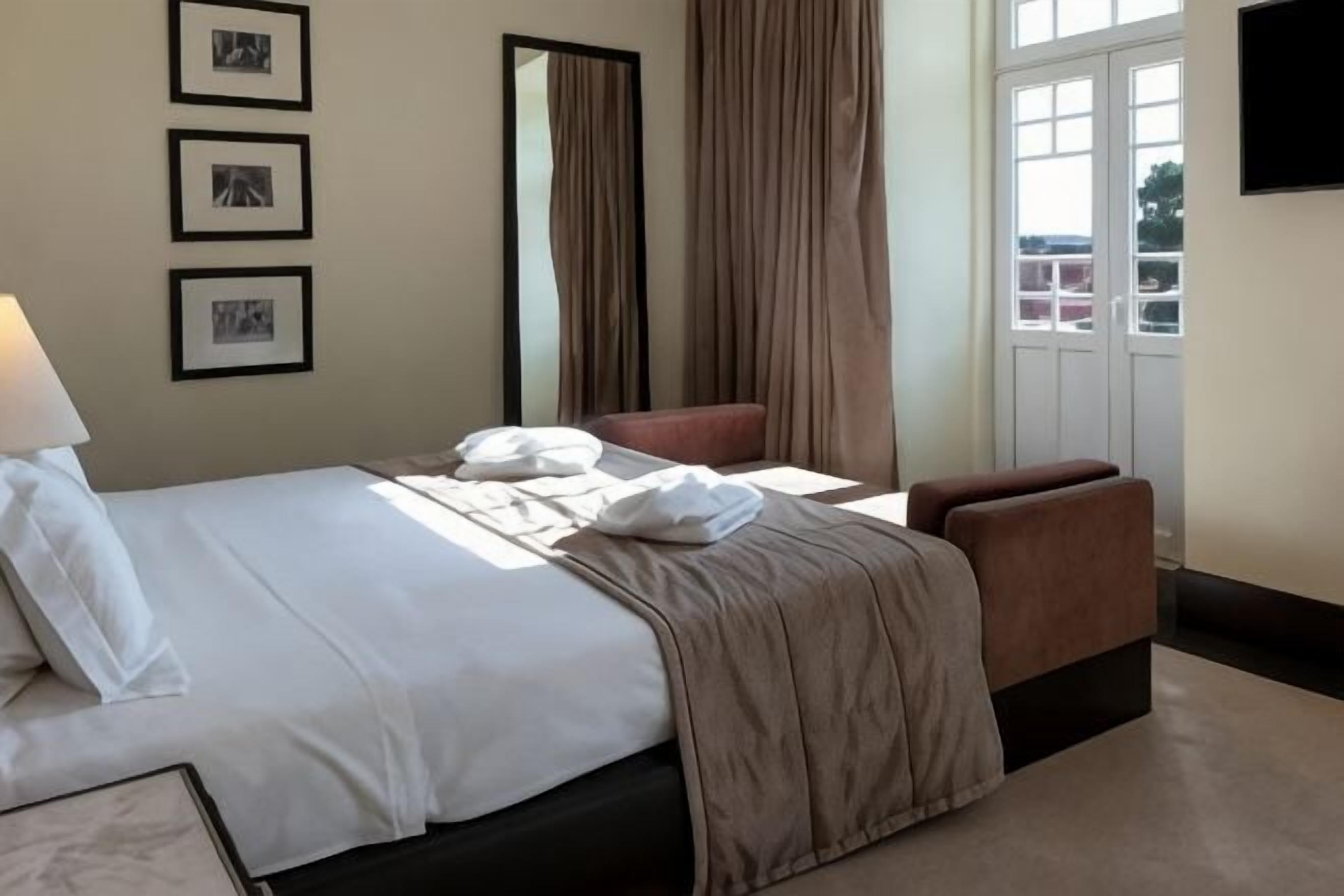 A hotel room with a bed, a chair, a desk, a mirror, and a window with curtains.