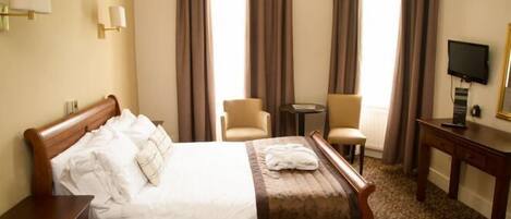 A hotel room with a wooden bed, two chairs, a small table, a TV, and curtains.