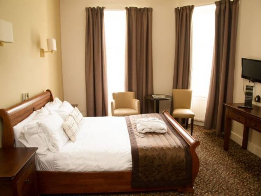 Ein Hotelzimmer mit einem hölzernen Bett, zwei Stühlen, einem kleinen Tisch, einem Fernseher und Vorhängen.