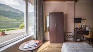 A hotel room with a bed, a desk, a chair, a wardrobe, and a window with a view of a lake and mountains.