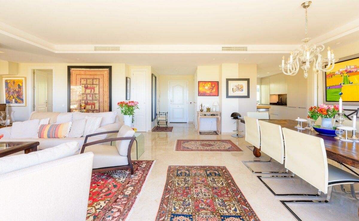 A spacious living room with a sofa set, a dining area with a long table, and a kitchen in the background.