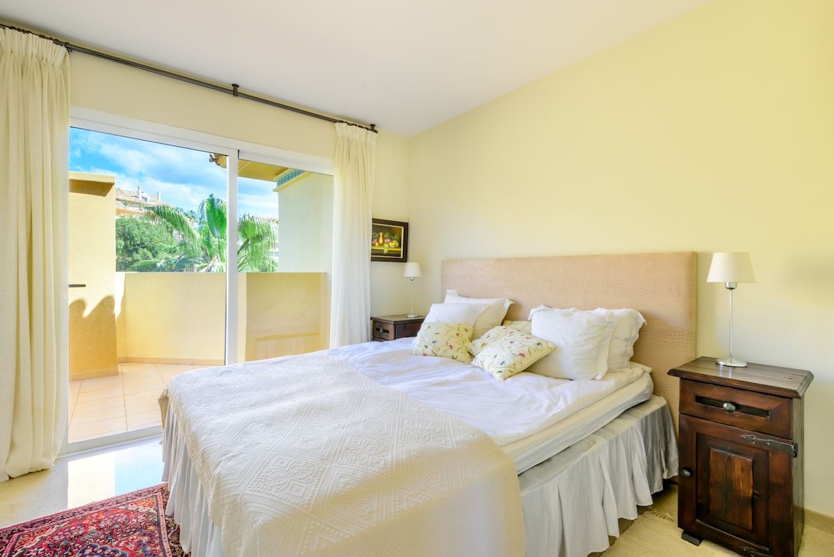 A bedroom with a bed, a nightstand, a lamp, and a view of the outdoors.