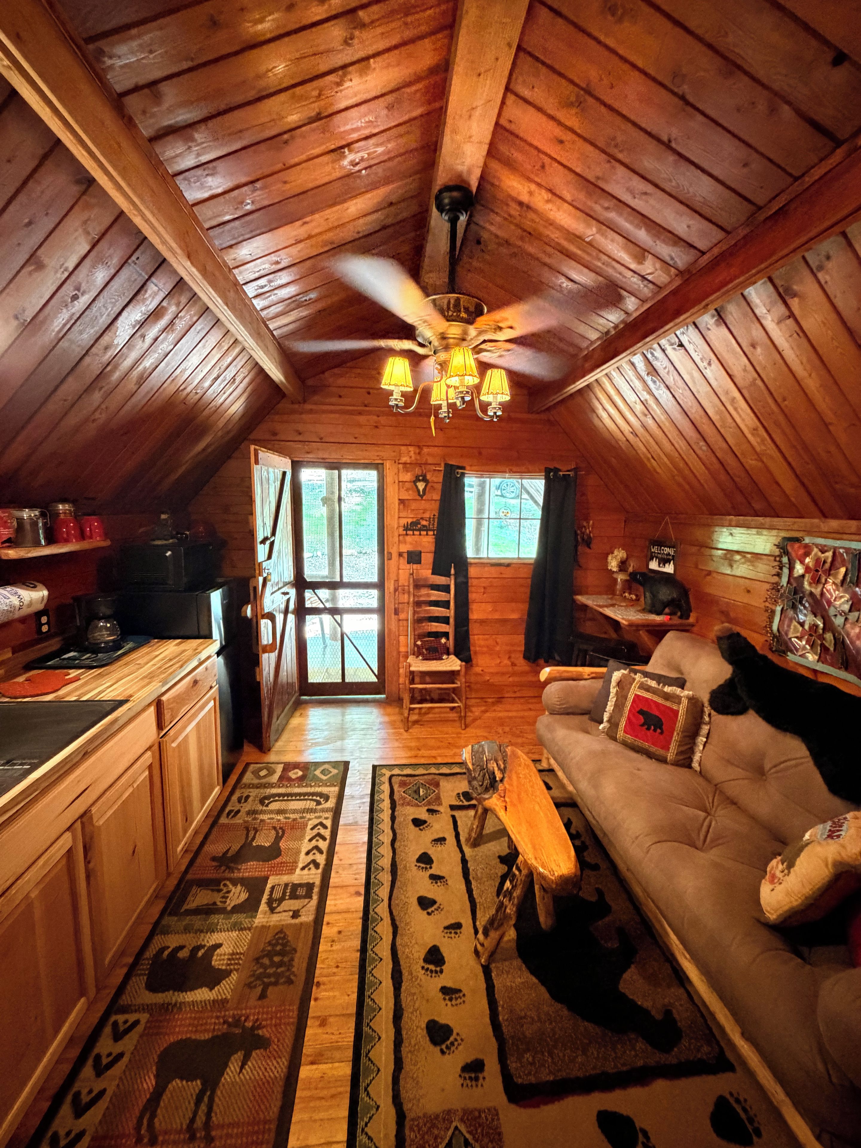 A cozy attic room with wooden beams, a ceiling fan, a sofa with a bear pillow, and a coffee table.