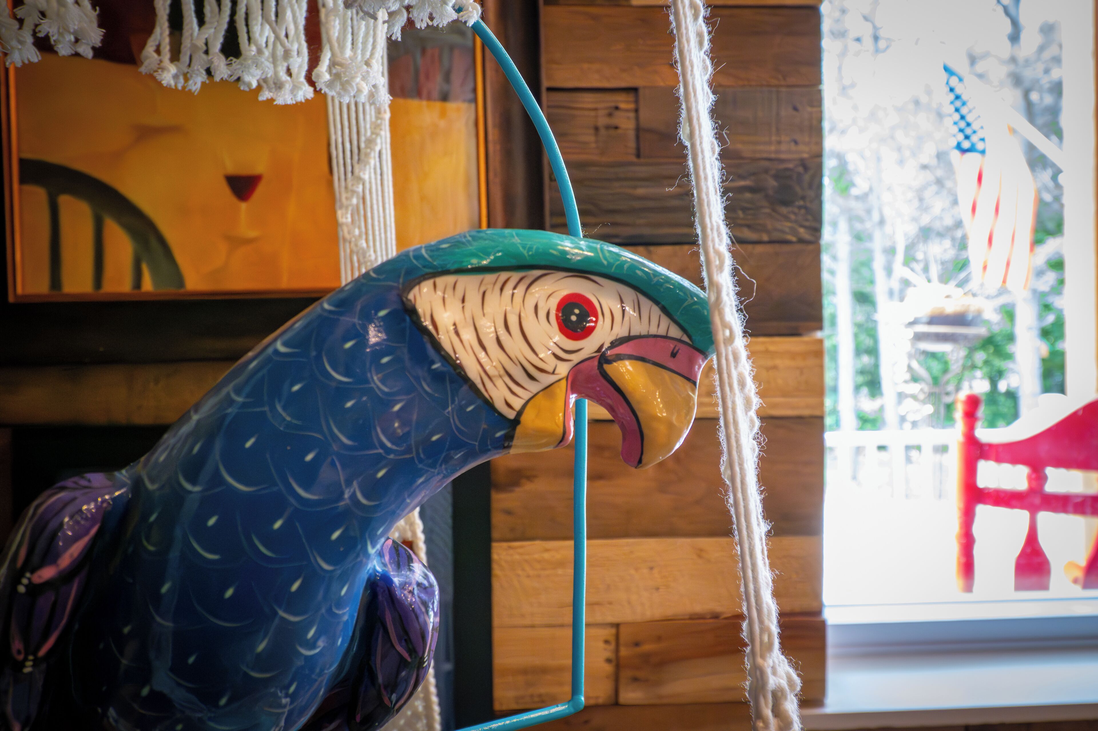 A colorful parrot sculpture in a room with wooden walls and a window with a view of a red chair and American flags.