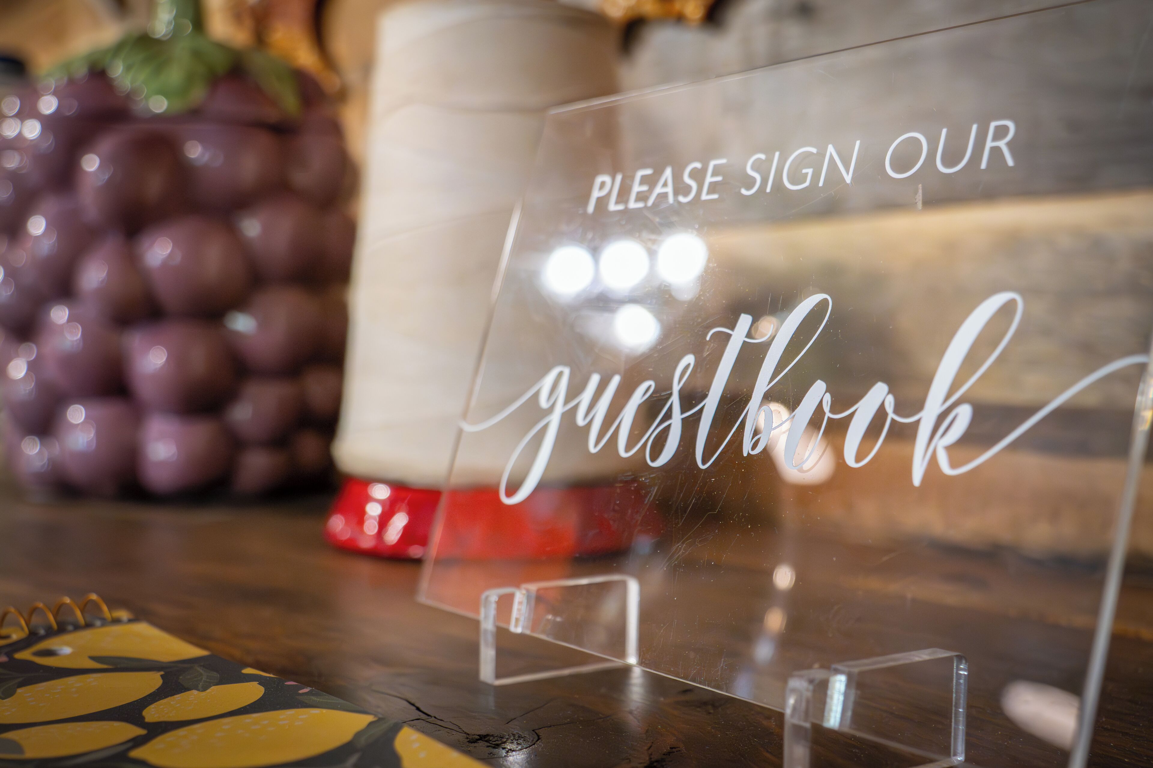 Un panneau transparent en acrylique avec l’inscription « VEUILLEZ INSCRIRE VOTRE NOM DANS NOTRE LIVRE D’OR », posé sur une table en bois.