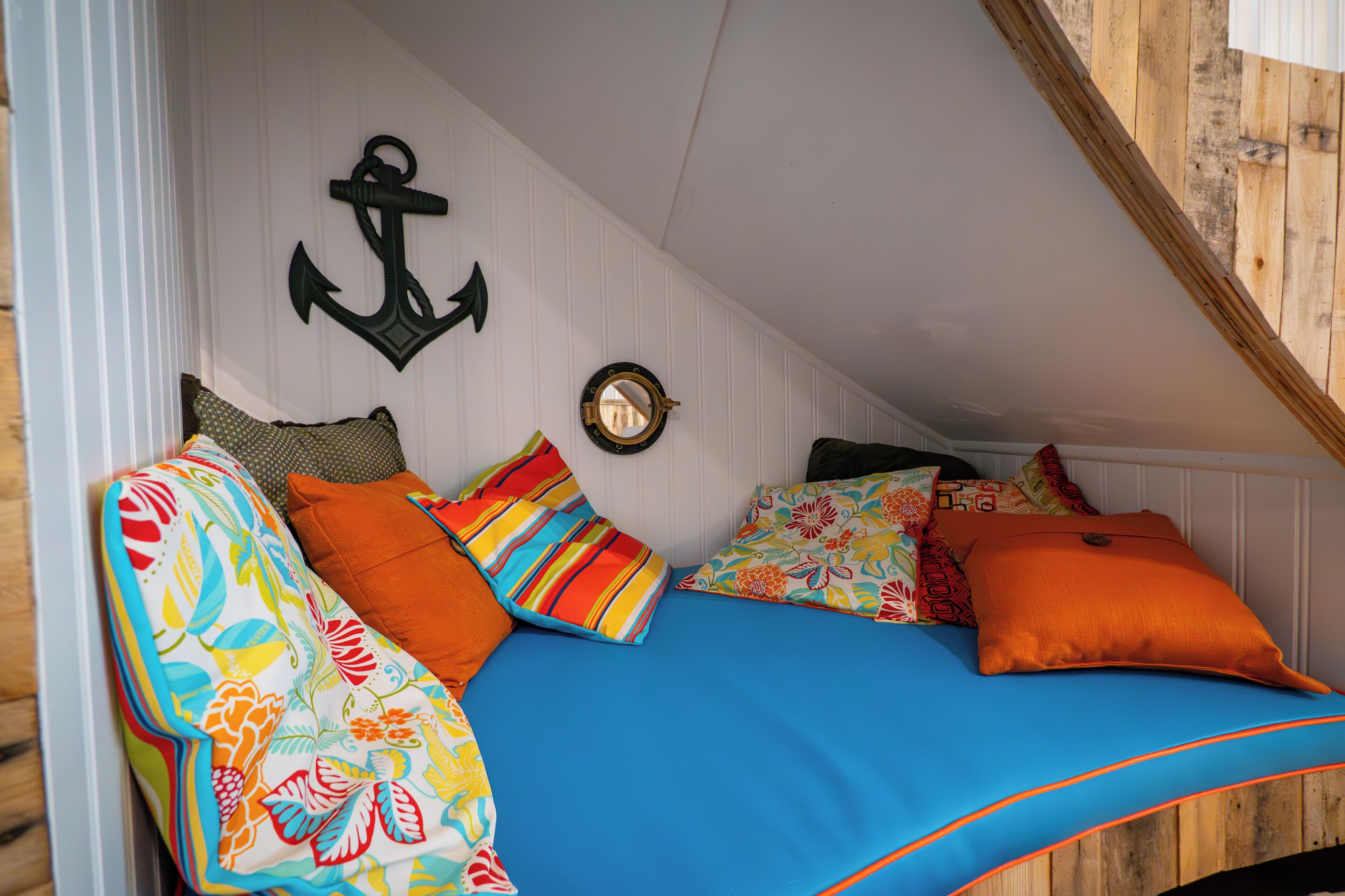 Une chambre mansardée chaleureuse, avec un plafond en pente, un lit recouvert d’une literie aux couleurs vives et une décoration sur le thème de la marine, comprenant une ancre et un miroir rond.