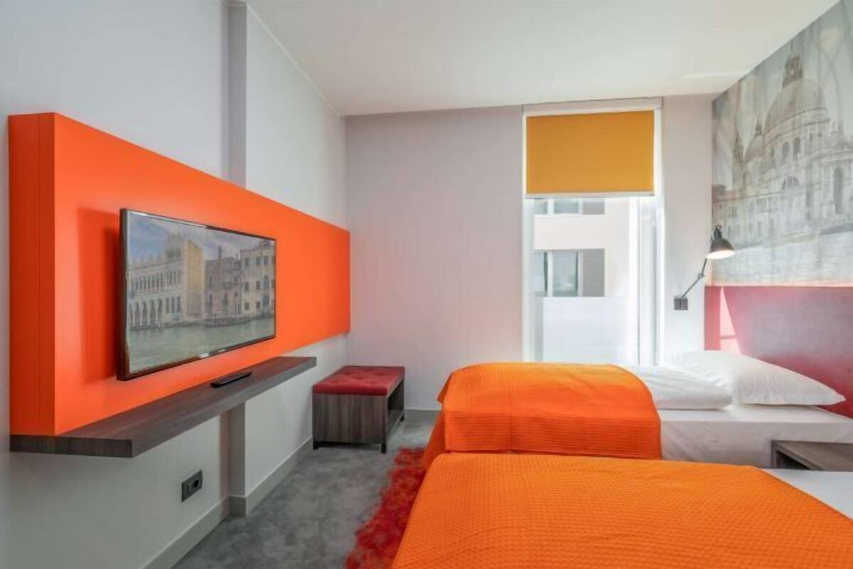 A hotel room with two beds, a TV mounted on a bright orange wall, and a painting of a building on the wall.