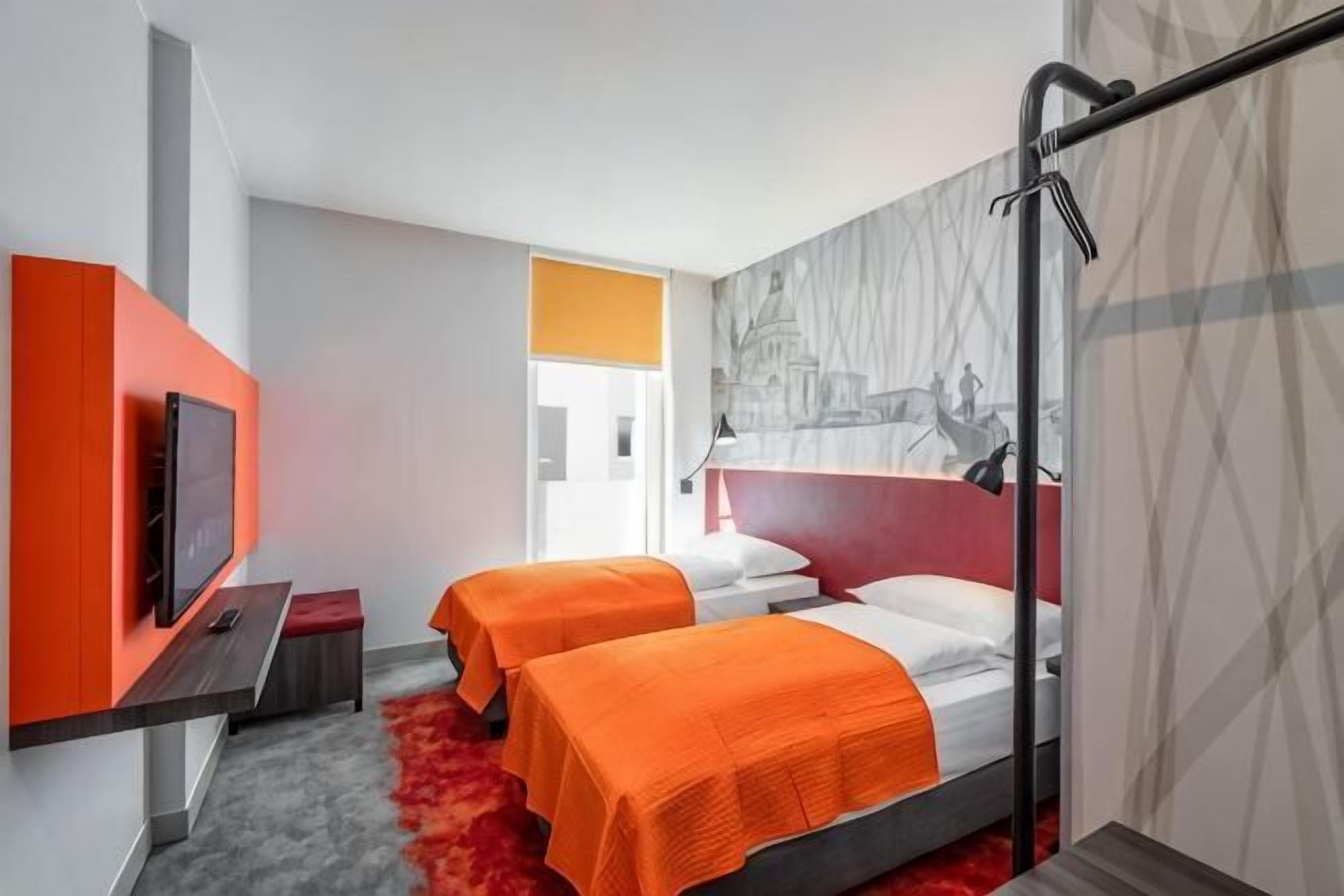 A hotel room with two beds, a TV, a red carpet, and a cityscape mural.