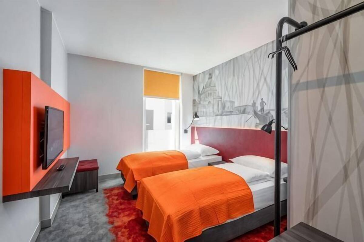 A hotel room with two beds, a TV, a red carpet, and a cityscape mural.