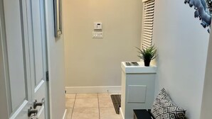Un couloir avec une porte à gauche, un banc avec des coussins à motifs à droite, une petite table avec une plante et un climatiseur fixé au mur.