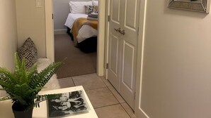 Un couloir avec un banc blanc, une plante en pot et un tableau encadré. Une porte donne sur une chambre à coucher qui contient un lit et une commode.