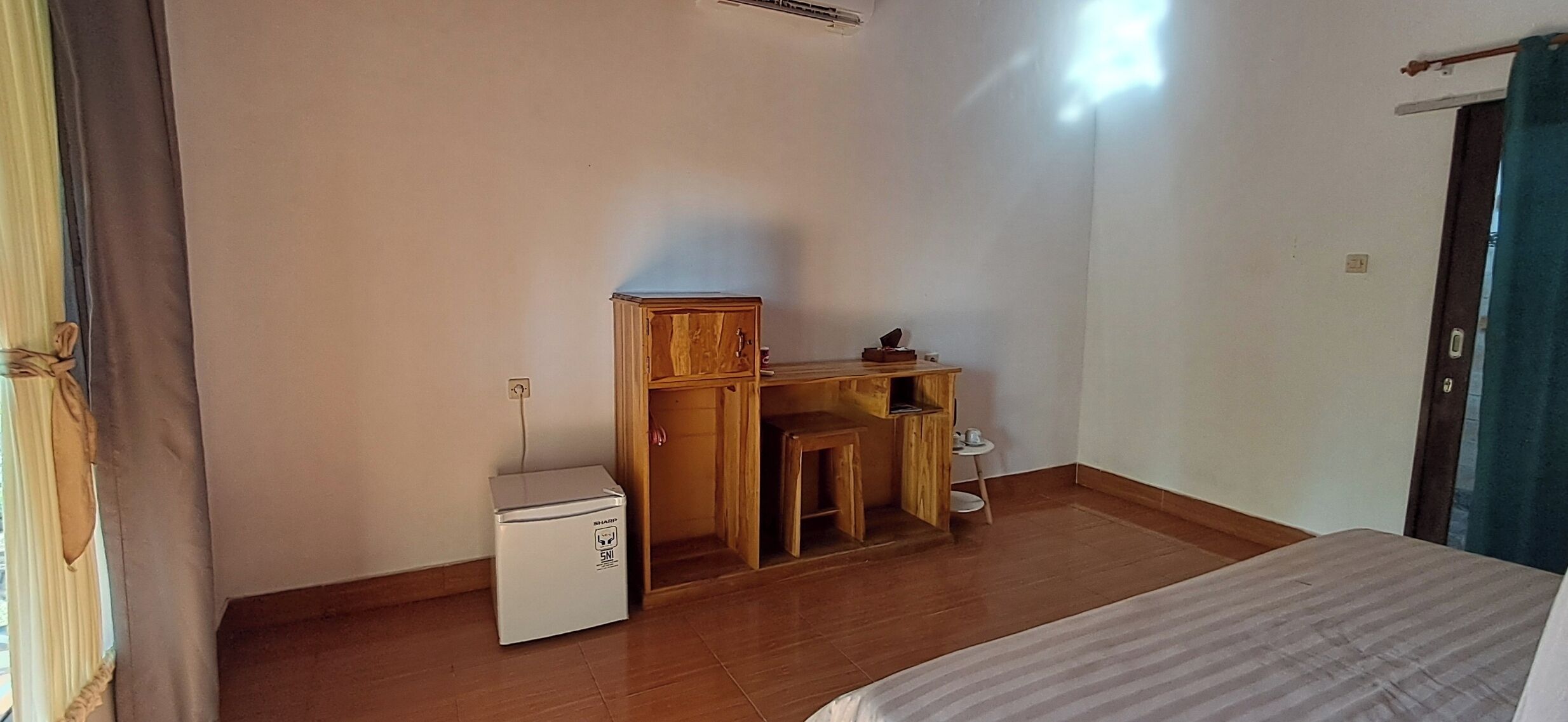 A small, simple room with a bed, a wooden desk, a small refrigerator, and a window with curtains.