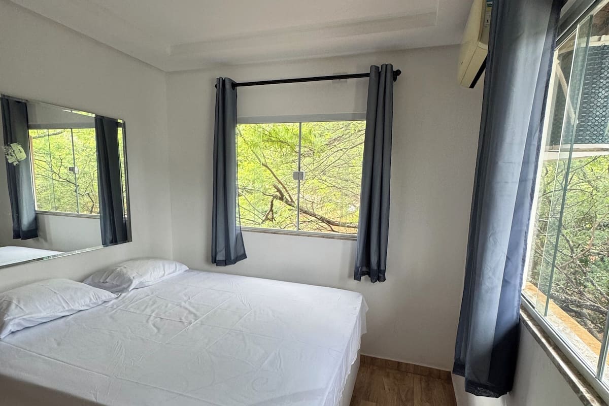 Ein Schlafzimmer mit einem großen Bett, zwei mit Vorhängen versehenen Fenstern, einem Deckenventilator und Blick auf Bäume.