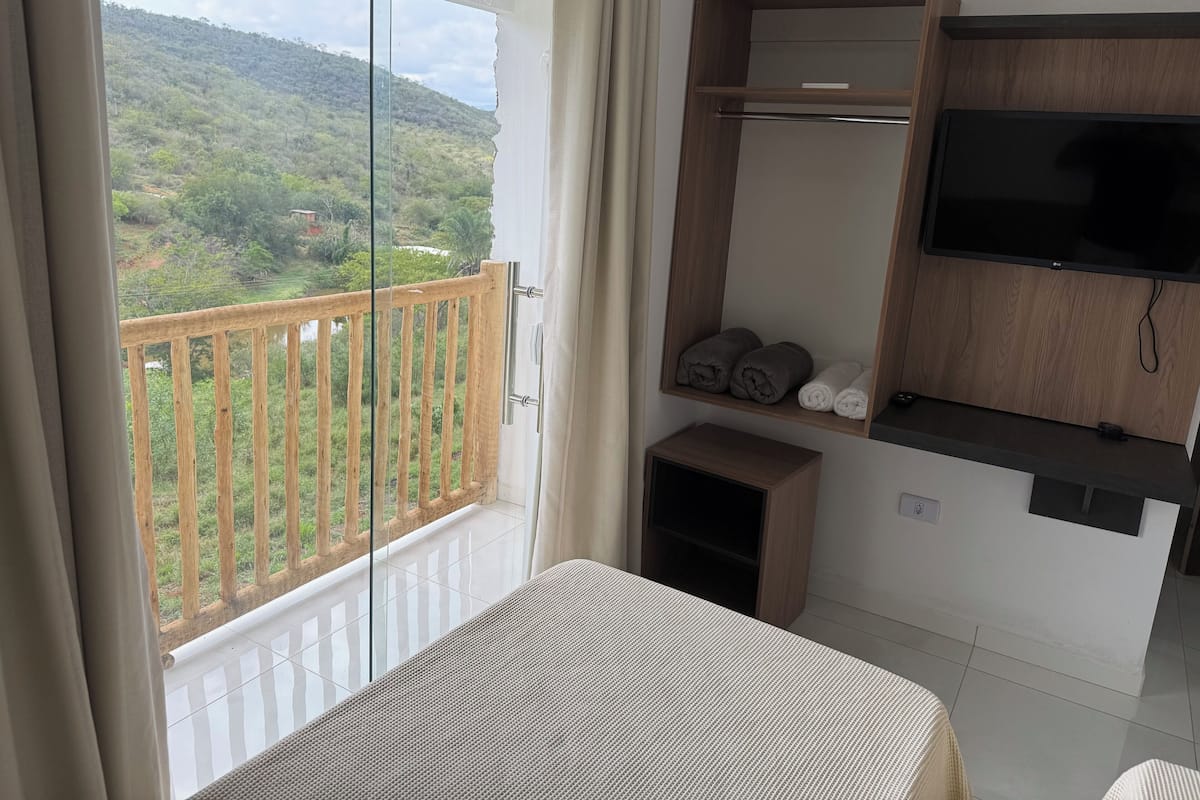 Ein Balkon mit Holzbalkon, ein Bett, ein an einer Holzwand befestigtes Fernsehgerät und Blick auf eine grüne Landschaft.