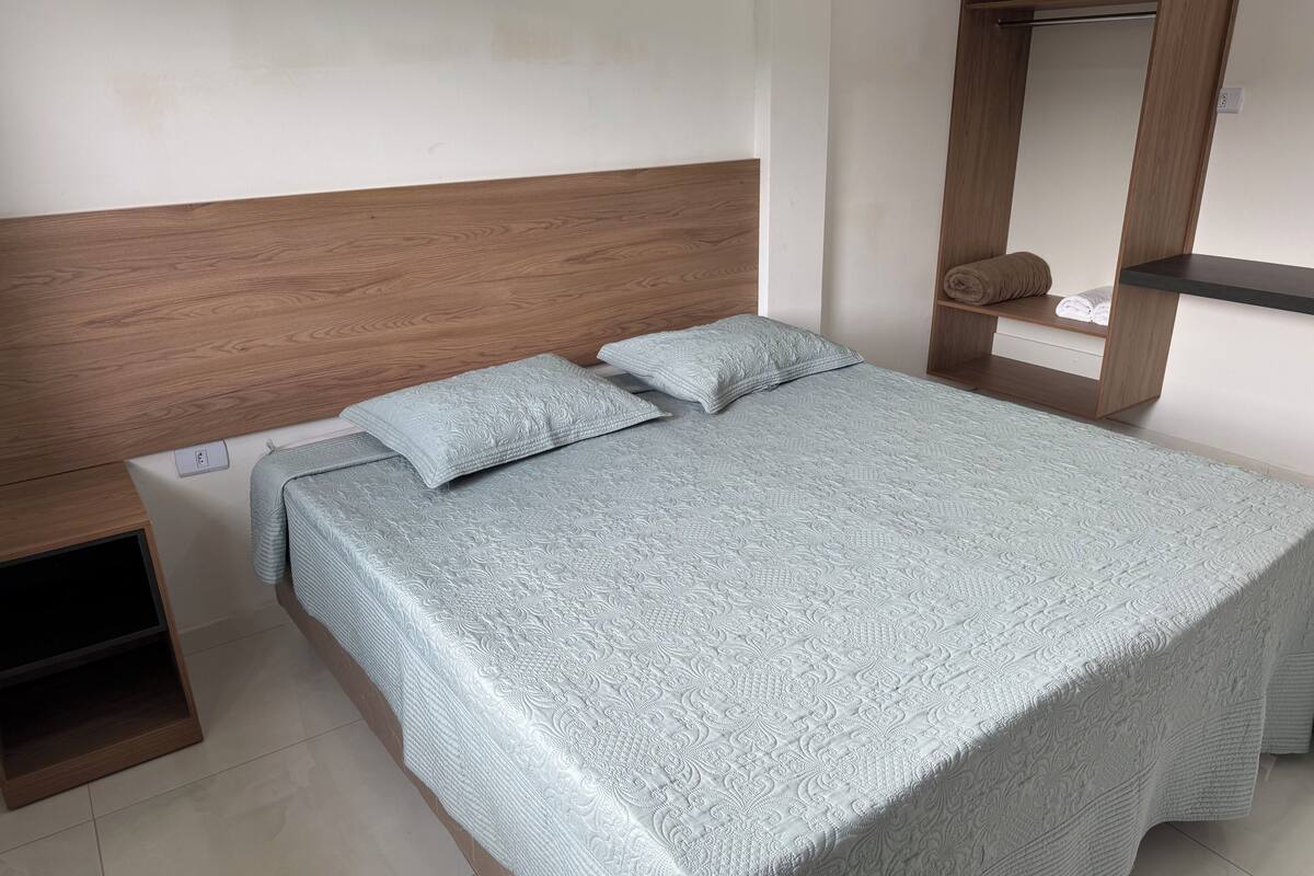 Ein ordentlich bezogenes Bett mit einem gemusterten Überwurf und zwei Kissen, ein Holz-Kopfteil, eine kleine Holztrennwand mit Handtüchern und ein Fliesenboden.