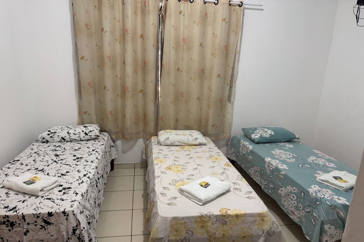 Ein kleines, sauberes Zimmer mit zwei Einzelbetten, jeweils mit einer blumigen Tagesdecke und einem Kopfkissen. Ein Vorhang mit Blumenmuster und ein kleiner Tisch mit einem Handtuch sind ebenfalls vorhanden.