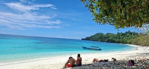 A beach with clear turquoise water, a boat, and people relaxing on the sand.