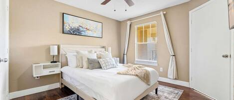 A bedroom with a bed, nightstands, a ceiling fan, a window with blinds, and a painting on the wall.