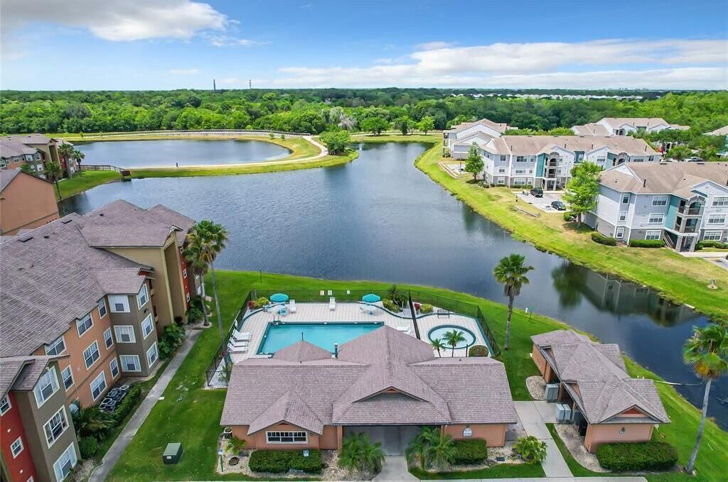 A residential area with a large swimming pool surrounded by apartment buildings, a lake, and lush greenery.