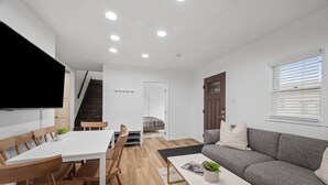 A modern living room with a white sofa, a dining table with wooden chairs, a TV mounted on the wall, and a staircase leading to another room. - Modern and Spacious 4 Bedroom Apt in Gibsonia/Pittsburgh (Bakerstown)