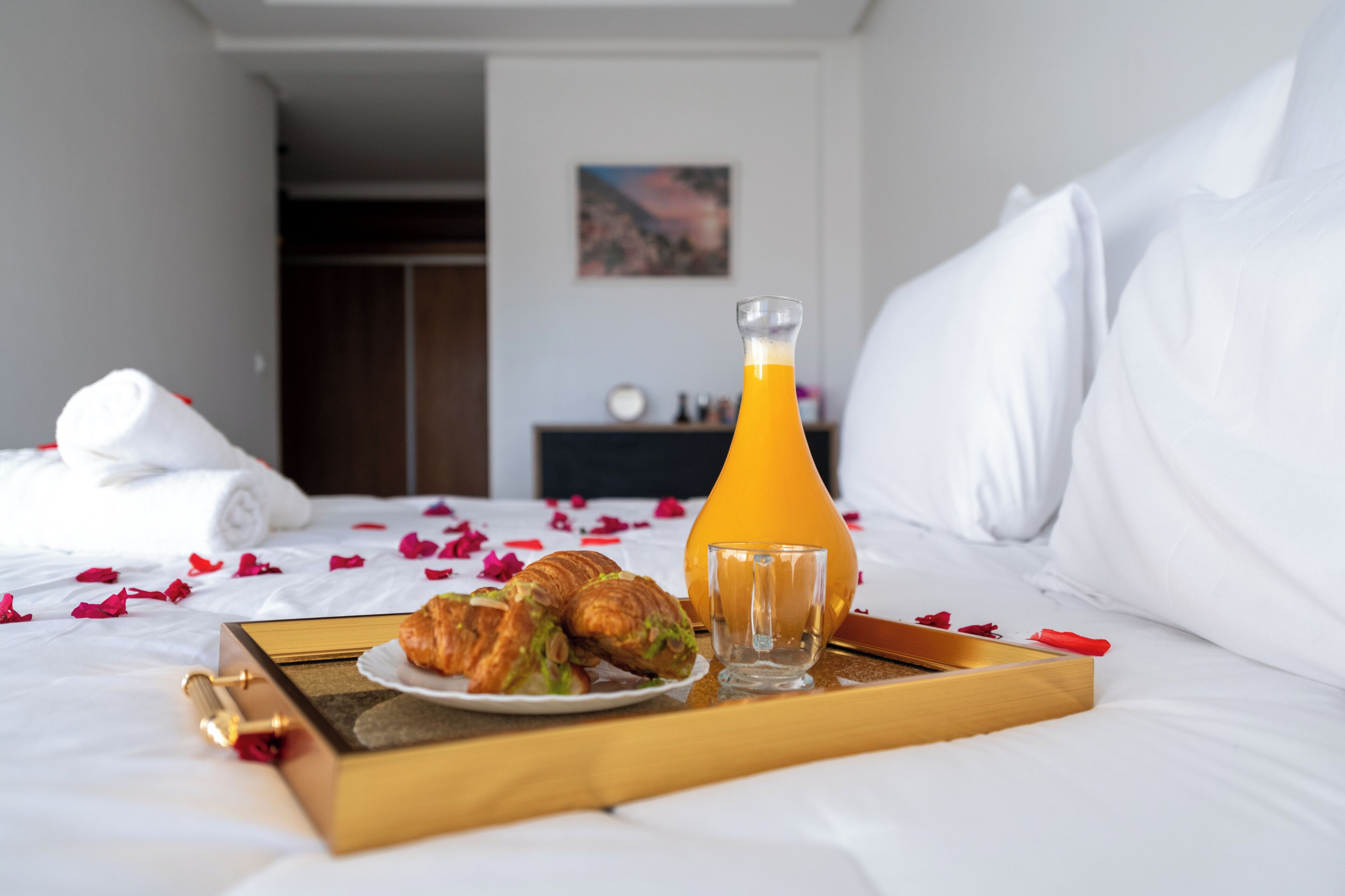 Un vassoio per la colazione con cornetti e un bicchiere di succo d'arancia su un letto con biancheria bianca e petali di rosa sparsi.
