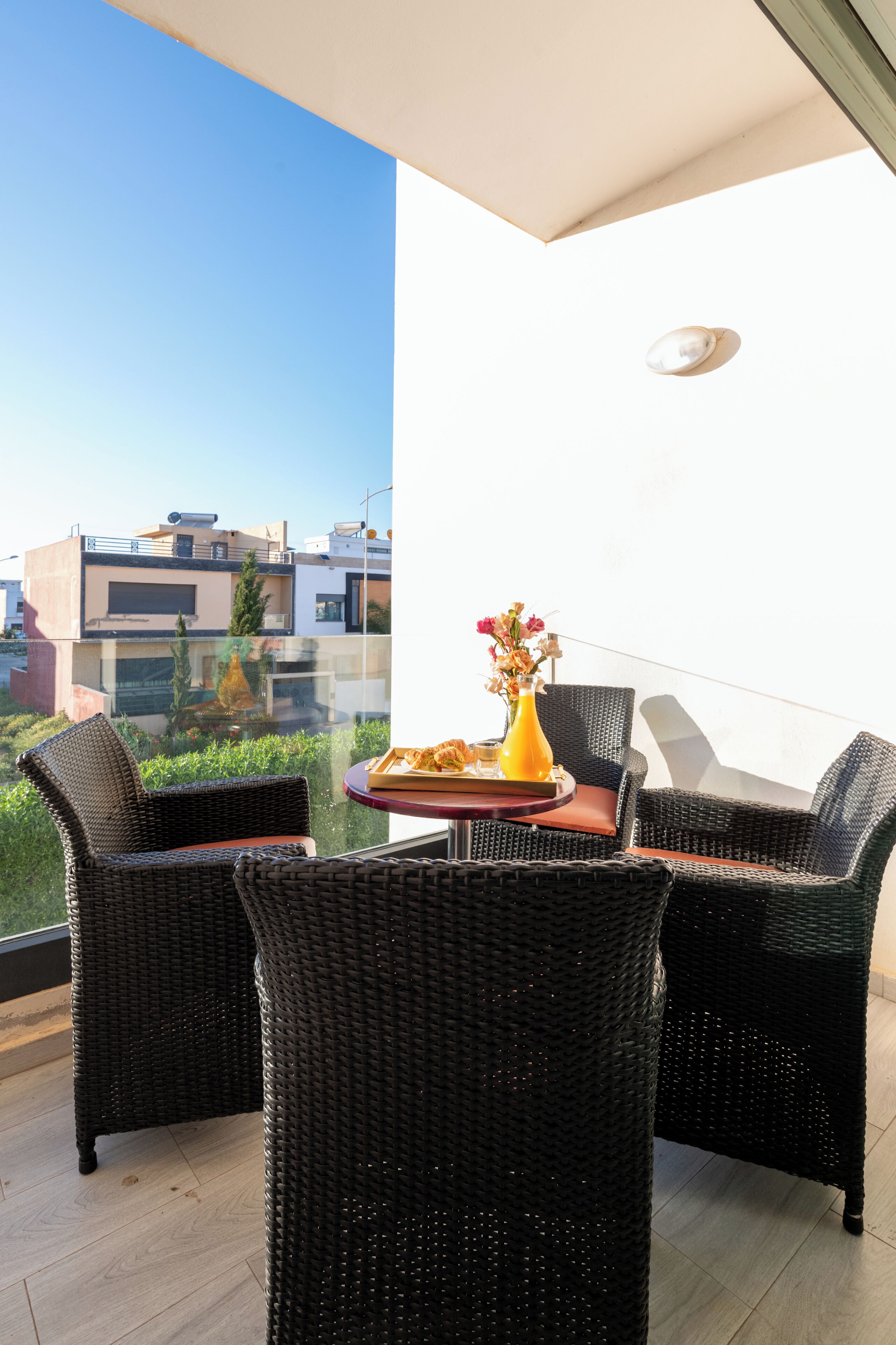 Een balkon met rieten meubilair, een tafel met een vaas en een schaal eten, en uitzicht op de stad.