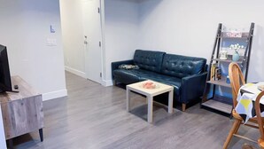Una sala de estar moderna con un sofá azul oscuro, una mesita blanca pequeña y una silla de madera. Un televisor está sobre un mueble de madera, y hay una estantería contra la pared.