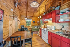 A wooden cabin interior with a dining table, chairs, a sofa, a kitchen with red cabinets, and a stove.