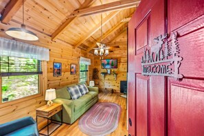 A cozy cabin interior with a green sofa, a small table with a lamp, and a red door with a welcome sign.