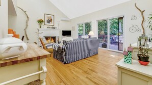 A spacious living room with a fireplace, a striped sofa, and a large window offering a view of the outdoors.