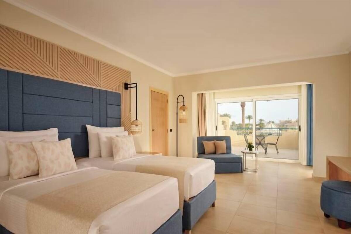 Habitación de hotel con dos camas, un balcón con vistas y una pequeña zona de estar.