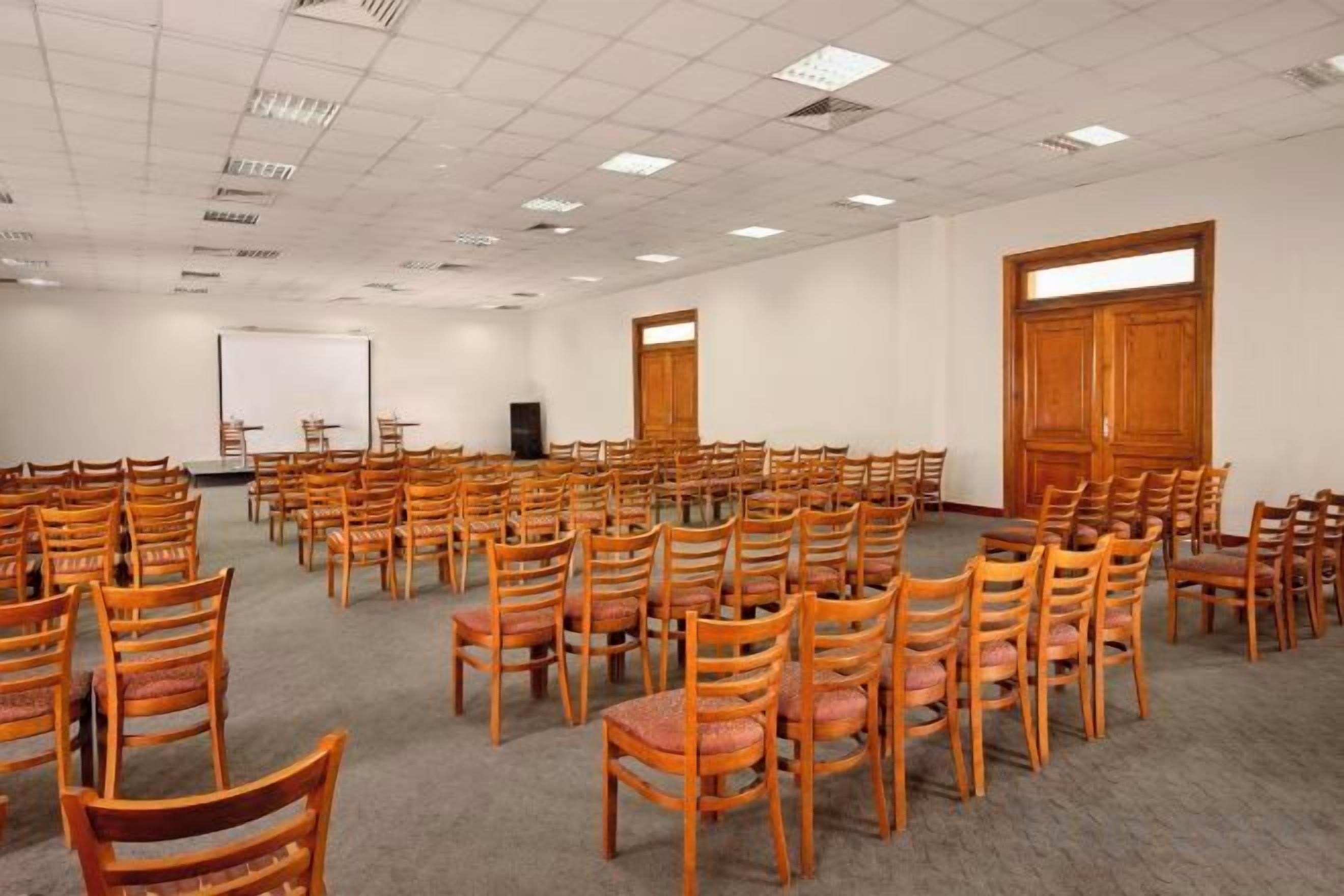 A conference room with rows of wooden chairs, a projector screen, and a large wooden door.