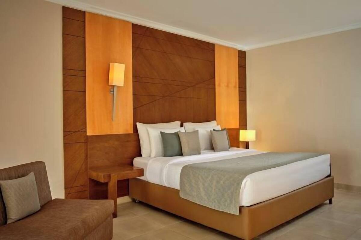 Una habitación de hotel moderna con una cama grande, cabecera de madera, dos mesitas de noche y un sillón marrón.